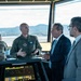 The image shows what appears to be a military air traffic control tower briefing, where a U.S. Air Force officer in a flight suit is speaking with two civilian officials in business attire. A female soldier in camouflage uniform is visible in the background working at the control station. The setting features large panoramic windows overlooking an airfield with mountains visible in the distance.