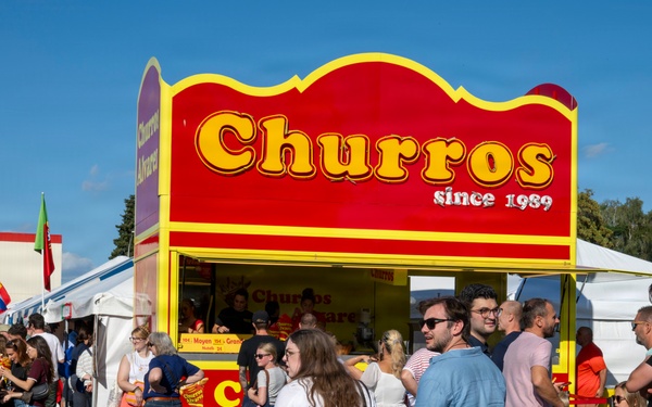 A busy outdoor food festival or fair featuring a prominent red and yellow churros stand labeled 'Churros since 1989.' A large crowd of people is gathered around the stall, many appearing to queue or socialize. The event takes place on a sunny day in what appears to be a parking lot or fairground area with white tents and international flags visible in the background.