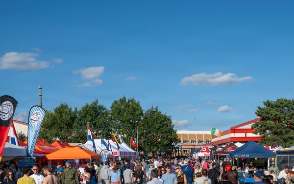 A large outdoor festival or fair is taking place in what appears to be a parking lot or open paved area. Multiple vendor tents, promotional banners, and national flags are visible, with a large crowd of attendees including families with strollers. The event appears to be a multicultural or international festival based on the variety of flags and vendor setups visible.