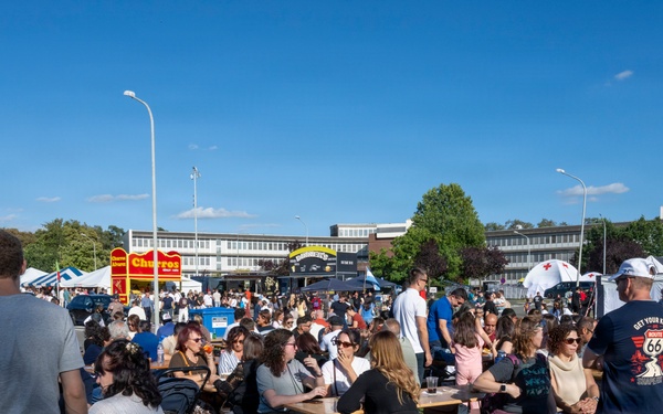 A large outdoor festival or food fair is taking place on a sunny day with clear blue skies. Crowds of people are seated at wooden picnic-style benches and tables on a grassy area, eating, drinking, and socializing. In the background, food vendor stalls including a visible 'Churros' stand, white tents, trees, and institutional buildings are visible.
