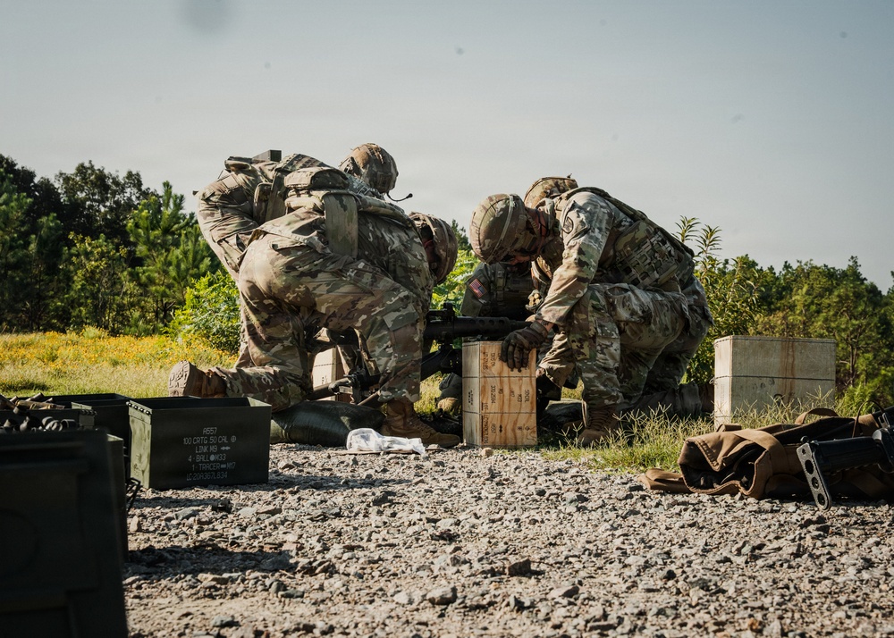 Two U.S. Army soldiers in Operational Camouflage Pattern (OCP) uniforms are working with a weapon system positioned on a wooden ammunition crate at what appears to be a military training range. Multiple ammunition cans and wooden crates are visible in the foreground and background. The setting is an outdoor gravel range with vegetation and trees in the background.