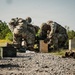 Two U.S. Army soldiers in Operational Camouflage Pattern (OCP) uniforms are working with a weapon system positioned on a wooden ammunition crate at what appears to be a military training range. Multiple ammunition cans and wooden crates are visible in the foreground and background. The setting is an outdoor gravel range with vegetation and trees in the background.
