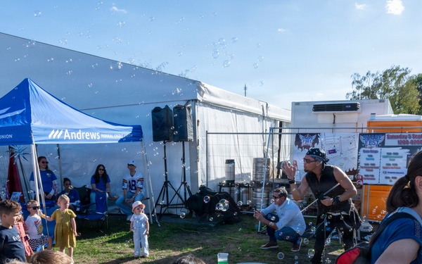 An outdoor family festival featuring a bubble performer entertaining a crowd of children and adults. A blue 'Andrews Federal Credit Union' branded canopy tent is prominently visible as a sponsor booth. The event appears to be a community fair held on a sunny day with multiple vendor tents and signage in the background.