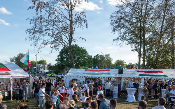 A large outdoor festival or cultural fair is taking place in an open park or fairground area. Multiple vendor tents are visible, decorated with what appear to be Bulgarian and Hungarian flags, suggesting a multicultural or international festival. A large crowd of attendees is gathered, watching what appears to be a performance or demonstration in the central open area.