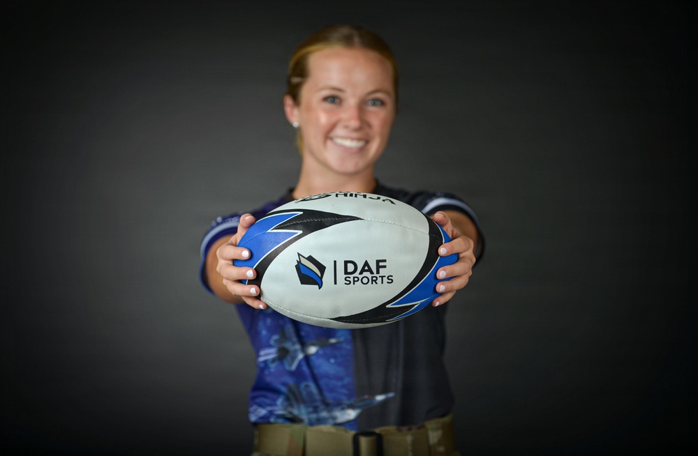 A smiling young woman holds a rugby ball toward the camera in a professional portrait-style photo. The ball prominently displays the 'DAF Sports' logo and branding. She is wearing a blue athletic jersey and appears to be in a studio setting with a dark background.