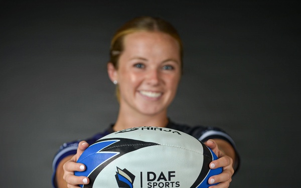 A smiling young woman holds a rugby ball toward the camera in a professional portrait-style photo. The ball prominently displays the 'DAF Sports' logo and branding. She is wearing a blue athletic jersey and appears to be in a studio setting with a dark background.