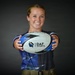 A smiling young woman holds a rugby ball toward the camera in a professional portrait-style photo. The ball prominently displays the 'DAF Sports' logo and branding. She is wearing a blue athletic jersey and appears to be in a studio setting with a dark background.