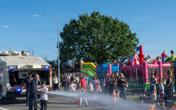 A public safety demonstration or open day event where firefighters or emergency personnel are allowing a child to operate a water hose connected to an emergency vehicle. The event appears festive, with colorful inflatable bounce houses visible in the background and a crowd of spectators watching. Orange traffic cones and blue barrier tape delineate the demonstration area on a wet road surface.
