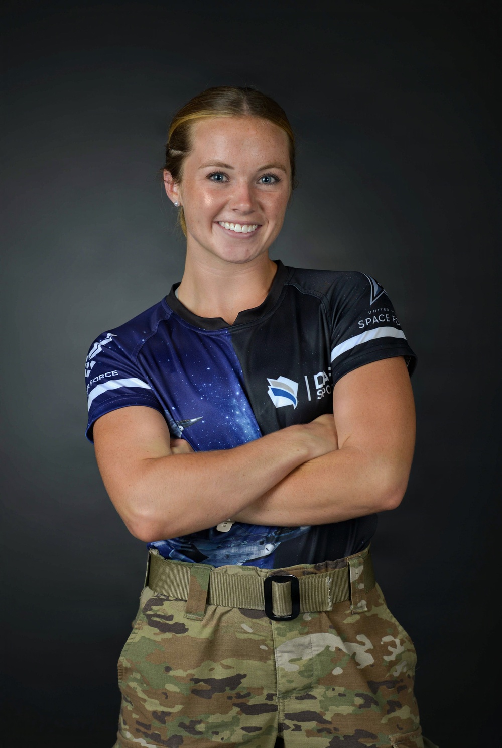 A portrait photograph of a person wearing a United States Space Force athletic jersey combined with military camouflage (MultiCam) trousers and a tan rigger's belt. The subject is posed with arms crossed and smiling against a dark studio background. The image appears to be an official or semi-official military sports portrait.