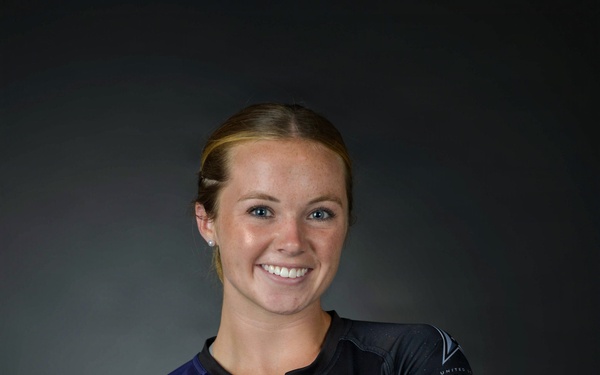 A portrait photograph of a person wearing a United States Space Force athletic jersey combined with military camouflage (MultiCam) trousers and a tan rigger's belt. The subject is posed with arms crossed and smiling against a dark studio background. The image appears to be an official or semi-official military sports portrait.