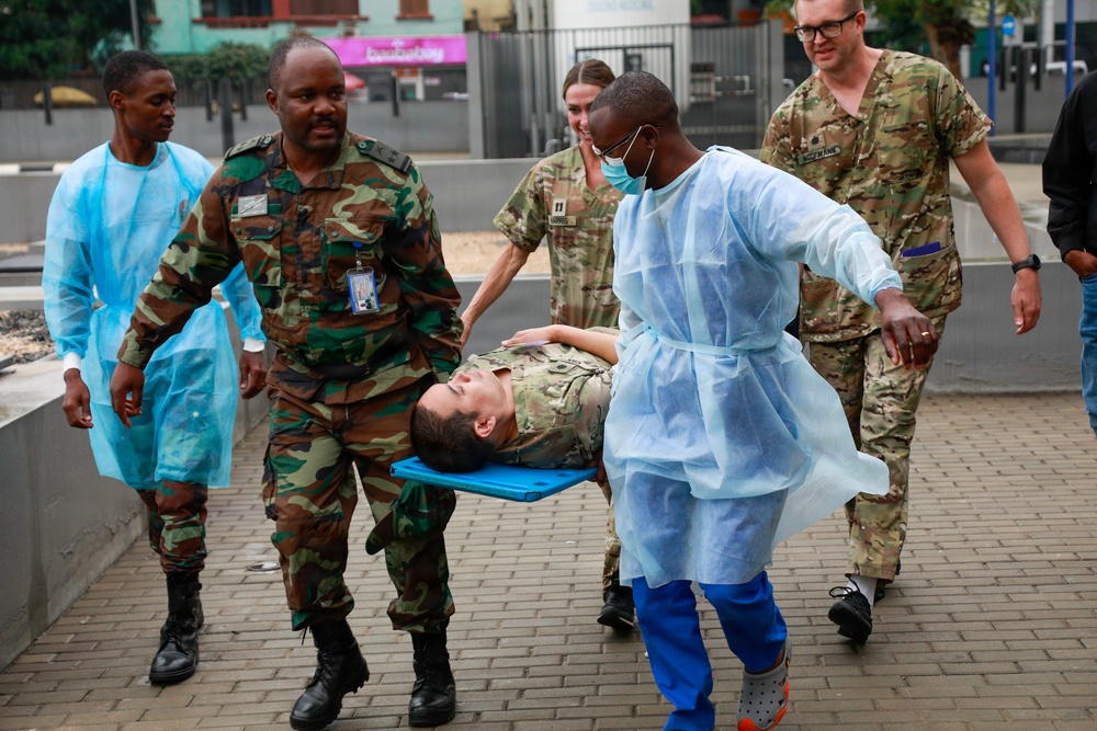 A group of military and medical personnel are carrying a person on a stretcher across a brick-paved outdoor area. The scene appears to depict a medical evacuation drill or training exercise, involving both military uniformed individuals and personnel wearing disposable medical gowns. Multiple nationalities of military uniforms are visible, suggesting a joint or multinational training exercise.