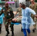A group of military and medical personnel are carrying a person on a stretcher across a brick-paved outdoor area. The scene appears to depict a medical evacuation drill or training exercise, involving both military uniformed individuals and personnel wearing disposable medical gowns. Multiple nationalities of military uniforms are visible, suggesting a joint or multinational training exercise.