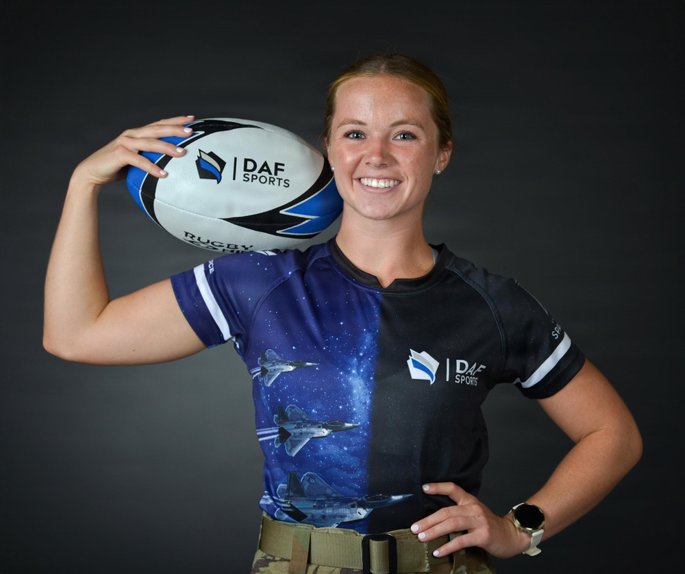 A young woman poses for a promotional sports portrait holding a rugby ball on her shoulder. She is wearing a DAF Sports jersey featuring a space/galaxy design with military aircraft graphics, paired with what appears to be military-style tan/khaki pants with a belt. The image appears to be an official sports team or promotional photograph.