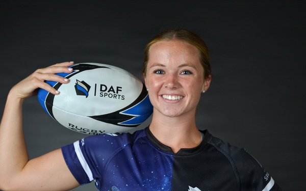 A young woman poses for a promotional sports portrait holding a rugby ball on her shoulder. She is wearing a DAF Sports jersey featuring a space/galaxy design with military aircraft graphics, paired with what appears to be military-style tan/khaki pants with a belt. The image appears to be an official sports team or promotional photograph.