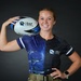 A young woman poses for a promotional sports portrait holding a rugby ball on her shoulder. She is wearing a DAF Sports jersey featuring a space/galaxy design with military aircraft graphics, paired with what appears to be military-style tan/khaki pants with a belt. The image appears to be an official sports team or promotional photograph.