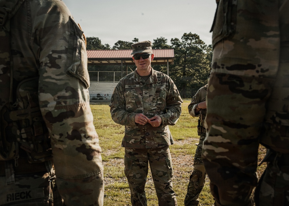 A U.S. Army soldier identified by name tape as 'MELTON' stands in the center of a group of soldiers in an outdoor field setting. He appears to be addressing or briefing the surrounding personnel, all wearing OCP (Operational Camouflage Pattern) uniforms. A covered bleacher structure with a red roof is visible in the background.