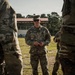 A U.S. Army soldier identified by name tape as 'MELTON' stands in the center of a group of soldiers in an outdoor field setting. He appears to be addressing or briefing the surrounding personnel, all wearing OCP (Operational Camouflage Pattern) uniforms. A covered bleacher structure with a red roof is visible in the background.