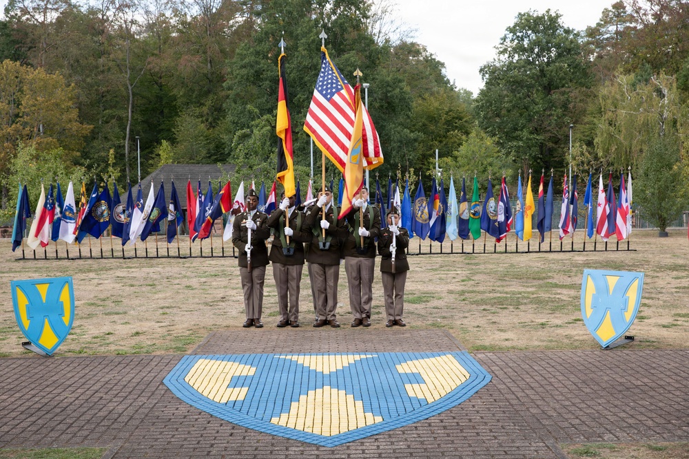 A military color guard ceremony featuring four uniformed soldiers holding flags, including the United States flag and the German flag, standing on a decorative brick plaza. The ceremony takes place in front of a semicircular display of numerous state and organizational flags mounted on a fence. The shield insignia visible on the ground and flanking displays appears to be associated with the U.S. Army's 21st Theater Sustainment Command (21st TSC), suggesting this is a military installation in Germany.