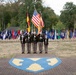 A military color guard ceremony featuring four uniformed soldiers holding flags, including the United States flag and the German flag, standing on a decorative brick plaza. The ceremony takes place in front of a semicircular display of numerous state and organizational flags mounted on a fence. The shield insignia visible on the ground and flanking displays appears to be associated with the U.S. Army's 21st Theater Sustainment Command (21st TSC), suggesting this is a military installation in Germany.