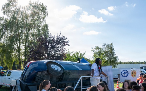 An outdoor public awareness event featuring an upside-down car mounted on a trailer, likely a road safety demonstration. A crowd of young people, possibly students or teenagers, are gathered around the display behind a blue rope barrier. The event appears to be taking place in a park or institutional grounds on a sunny day.
