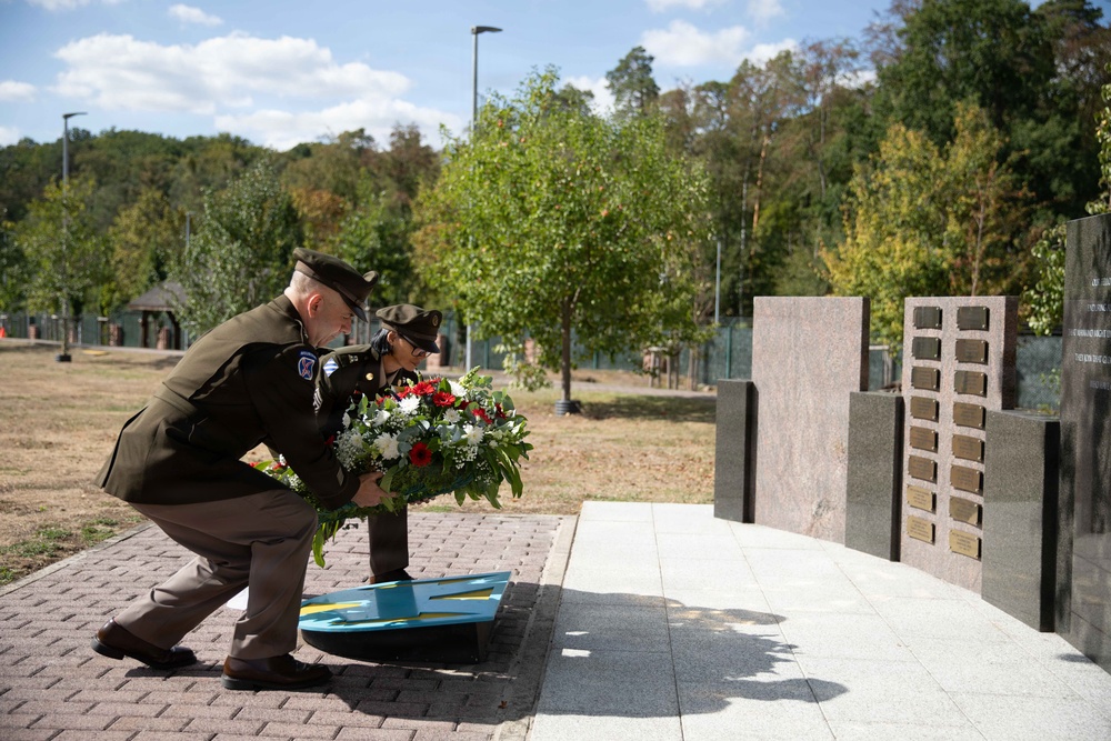 Two military personnel in olive drab uniforms are laying a floral wreath at a memorial monument during a formal ceremony. The memorial features dark granite walls with bronze plaques bearing names, and a decorative element in blue and yellow is visible at the base. The setting is an outdoor memorial park with trees and manicured grounds in the background.