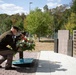 Two military personnel in olive drab uniforms are laying a floral wreath at a memorial monument during a formal ceremony. The memorial features dark granite walls with bronze plaques bearing names, and a decorative element in blue and yellow is visible at the base. The setting is an outdoor memorial park with trees and manicured grounds in the background.
