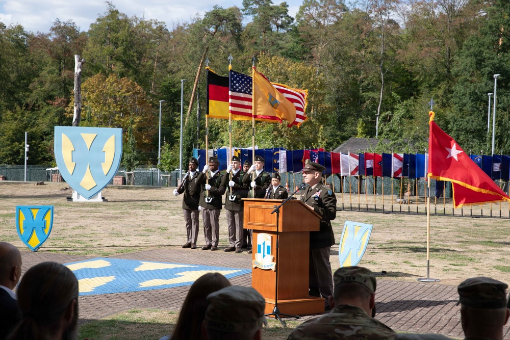 A formal U.S. Army military ceremony is taking place outdoors, likely at a base in Germany given the presence of a German flag alongside the U.S. flag. A uniformed officer is speaking at a wooden podium while a color guard stands at attention behind him holding multiple flags. The setting features unit insignia displays, state flags, and a one-star general's flag, suggesting a change of command or activation ceremony.