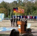A formal U.S. Army military ceremony is taking place outdoors, likely at a base in Germany given the presence of a German flag alongside the U.S. flag. A uniformed officer is speaking at a wooden podium while a color guard stands at attention behind him holding multiple flags. The setting features unit insignia displays, state flags, and a one-star general's flag, suggesting a change of command or activation ceremony.