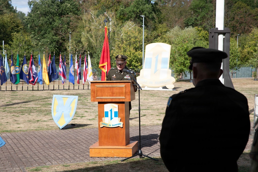 A U.S. Army officer in dress uniform delivers a speech at a formal military ceremony from a wooden podium bearing the '1st Theater Sustainment Command' insignia with the motto 'First in Support.' The event takes place outdoors on a brick plaza surrounded by multiple state and military flags, with a large unit monument visible in the background. A second uniformed figure stands in the foreground observing the proceedings.