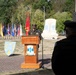 A U.S. Army officer in dress uniform delivers a speech at a formal military ceremony from a wooden podium bearing the '1st Theater Sustainment Command' insignia with the motto 'First in Support.' The event takes place outdoors on a brick plaza surrounded by multiple state and military flags, with a large unit monument visible in the background. A second uniformed figure stands in the foreground observing the proceedings.