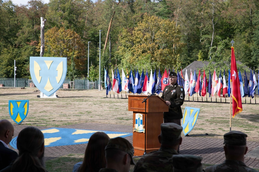A U.S. Army staff sergeant or senior NCO is delivering a speech at a formal military ceremony from a wooden podium. The event appears to be an official unit ceremony, likely a change of command or activation/inactivation ceremony, held outdoors on a military installation. Multiple state and unit flags are displayed in the background alongside large unit insignia displays.