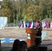 A U.S. Army staff sergeant or senior NCO is delivering a speech at a formal military ceremony from a wooden podium. The event appears to be an official unit ceremony, likely a change of command or activation/inactivation ceremony, held outdoors on a military installation. Multiple state and unit flags are displayed in the background alongside large unit insignia displays.