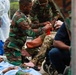 The image depicts what appears to be a mass casualty exercise or emergency medical response scenario involving military personnel and medical staff. Soldiers in camouflage uniforms are providing first aid to individuals on the ground and in a wheelchair, while personnel in white lab coats and scrubs assist nearby. The scene suggests a coordinated disaster response drill or real emergency triage situation.