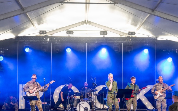 A military band performs on a lit stage under a white tent structure, with performers dressed in camouflage uniforms playing various instruments. A group of young children watches from behind a metal barrier in front of the stage, with some dancing and raising their hands. Two military personnel in uniform stand to the right side observing the performance.