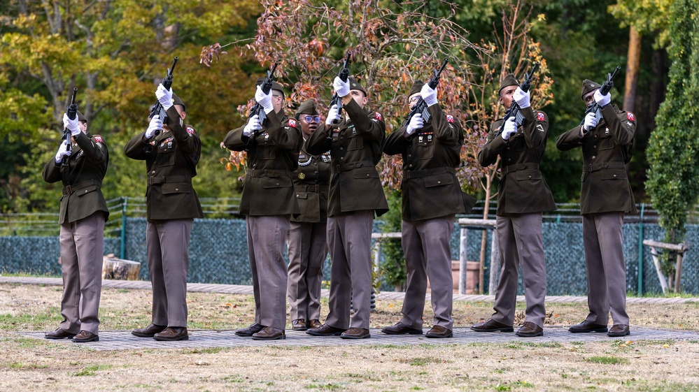 A military honor guard of seven soldiers in dress uniforms is performing a rifle volley salute, likely at a memorial or funeral ceremony. The soldiers are wearing olive/dark green dress jackets with gray trousers and white gloves, raising their rifles skyward in unison. The ceremony takes place outdoors on a paved platform surrounded by grass and autumn trees.