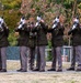 A military honor guard of seven soldiers in dress uniforms is performing a rifle volley salute, likely at a memorial or funeral ceremony. The soldiers are wearing olive/dark green dress jackets with gray trousers and white gloves, raising their rifles skyward in unison. The ceremony takes place outdoors on a paved platform surrounded by grass and autumn trees.