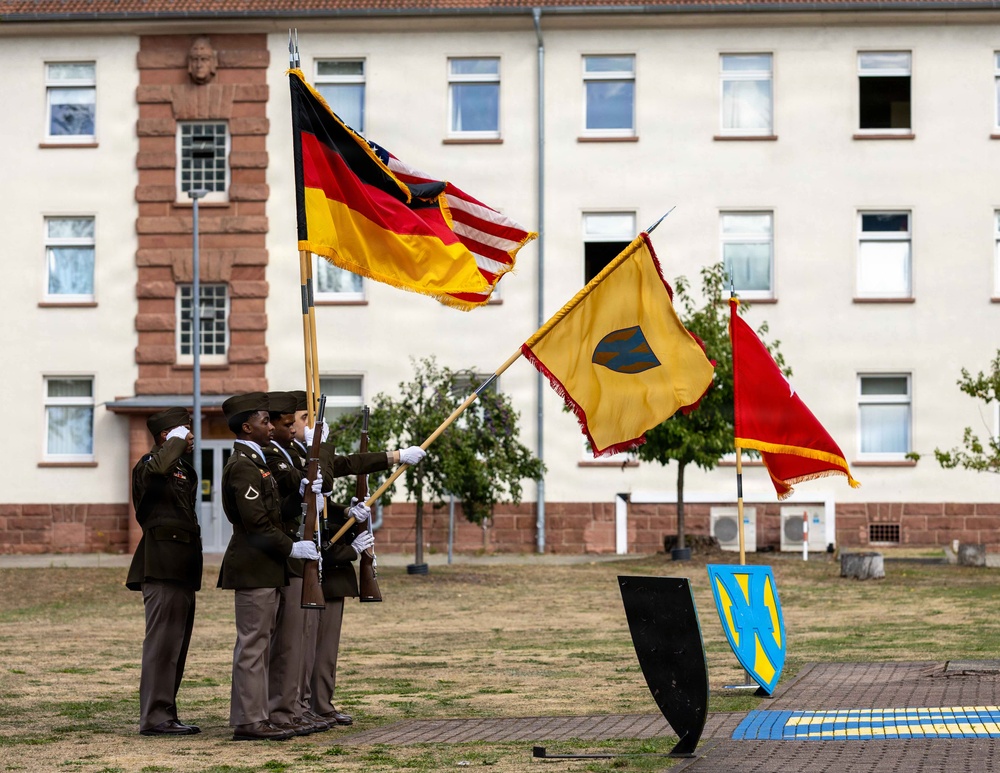 A U.S. Army color guard ceremony is taking place on what appears to be a military installation in Germany, evidenced by the German flag being carried alongside the U.S. flag. Soldiers in Army Service Uniforms are holding flags and rifles in a formal parade formation in front of a European-style military barracks building. A yellow unit guidon and a red flag with a star are also visible, along with decorative shield insignia on the ground.
