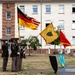 A U.S. Army color guard ceremony is taking place on what appears to be a military installation in Germany, evidenced by the German flag being carried alongside the U.S. flag. Soldiers in Army Service Uniforms are holding flags and rifles in a formal parade formation in front of a European-style military barracks building. A yellow unit guidon and a red flag with a star are also visible, along with decorative shield insignia on the ground.