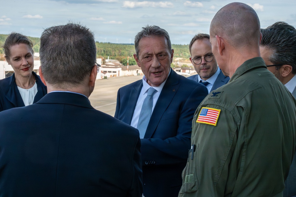 A group of civilians in formal business attire are meeting with a U.S. military officer in an olive green flight suit bearing an American flag patch on the shoulder. The gathering appears to take place on an airfield or military installation, visible in the background. The interaction appears to be an official diplomatic or liaison meeting.