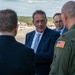 A group of civilians in formal business attire are meeting with a U.S. military officer in an olive green flight suit bearing an American flag patch on the shoulder. The gathering appears to take place on an airfield or military installation, visible in the background. The interaction appears to be an official diplomatic or liaison meeting.
