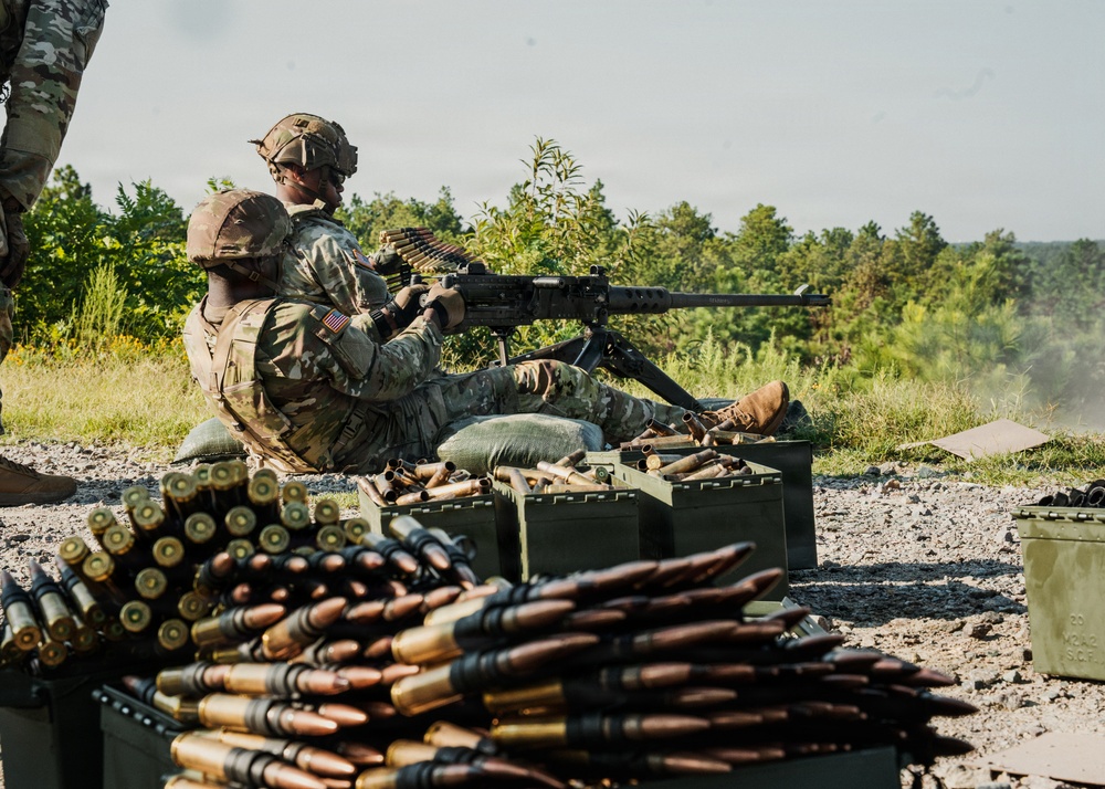 Two U.S. Army soldiers in MultiCam/OCP camouflage uniforms are operating a belt-fed machine gun from a prone/seated firing position on a gravel range. Large quantities of linked ammunition and green metal ammo cans are visible in the foreground. The setting appears to be an outdoor live-fire training range surrounded by trees and vegetation.