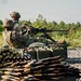 Two U.S. Army soldiers in MultiCam/OCP camouflage uniforms are operating a belt-fed machine gun from a prone/seated firing position on a gravel range. Large quantities of linked ammunition and green metal ammo cans are visible in the foreground. The setting appears to be an outdoor live-fire training range surrounded by trees and vegetation.
