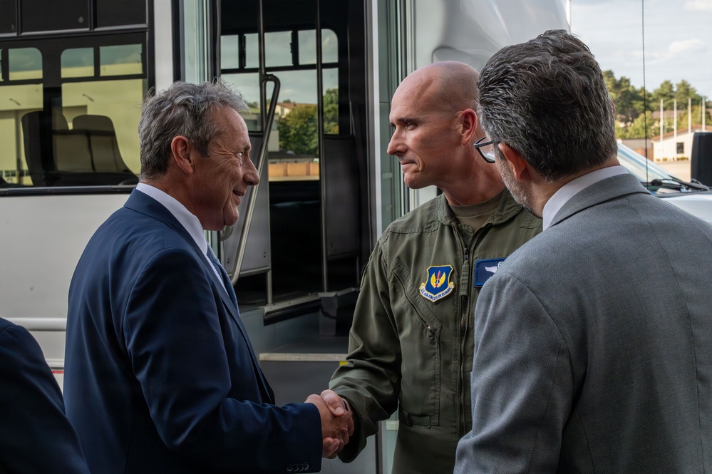 Three men are engaged in a formal greeting near what appears to be a military or government transport vehicle. A man in a blue suit is shaking hands with a military officer wearing an olive green flight suit bearing a U.S. Air Force in Europe patch. A third man in a gray suit stands nearby observing the exchange.