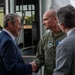 Three men are engaged in a formal greeting near what appears to be a military or government transport vehicle. A man in a blue suit is shaking hands with a military officer wearing an olive green flight suit bearing a U.S. Air Force in Europe patch. A third man in a gray suit stands nearby observing the exchange.
