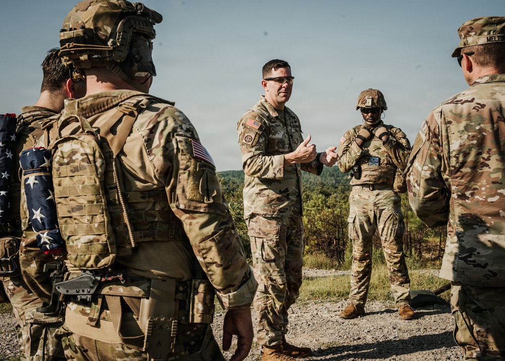 A group of U.S. Army soldiers in Operational Camouflage Pattern (OCP) uniforms are gathered outdoors in what appears to be a field training or briefing scenario. A central figure, wearing safety glasses and no helmet, appears to be addressing or debriefing the group with hand gestures. The setting is a gravel area with forested hills visible in the background.