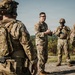 A group of U.S. Army soldiers in Operational Camouflage Pattern (OCP) uniforms are gathered outdoors in what appears to be a field training or briefing scenario. A central figure, wearing safety glasses and no helmet, appears to be addressing or debriefing the group with hand gestures. The setting is a gravel area with forested hills visible in the background.