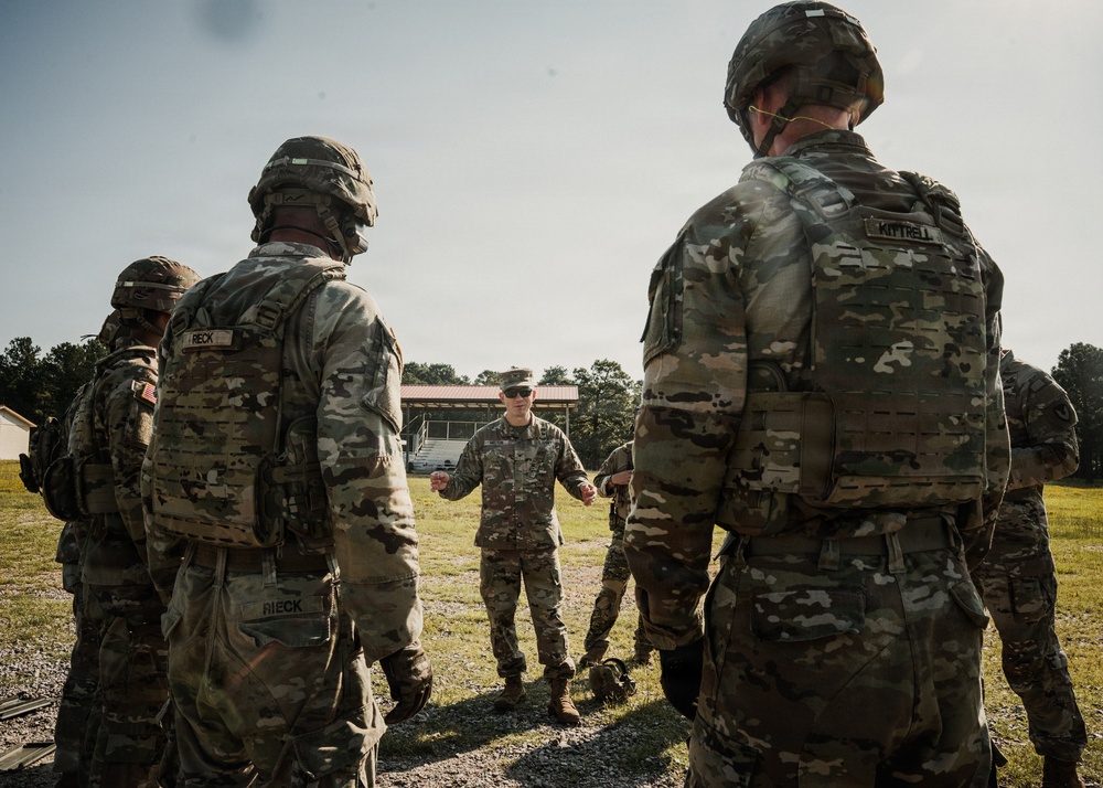 A group of U.S. Army soldiers in full combat gear, including OCP (Operational Camouflage Pattern) uniforms, helmets, and plate carriers, are gathered in a semi-circle outdoors. A central figure, wearing a patrol cap rather than a helmet, appears to be conducting a briefing or instruction session. The setting appears to be a military training area with a building and grassy field visible in the background.