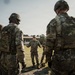 A group of U.S. Army soldiers in full combat gear, including OCP (Operational Camouflage Pattern) uniforms, helmets, and plate carriers, are gathered in a semi-circle outdoors. A central figure, wearing a patrol cap rather than a helmet, appears to be conducting a briefing or instruction session. The setting appears to be a military training area with a building and grassy field visible in the background.