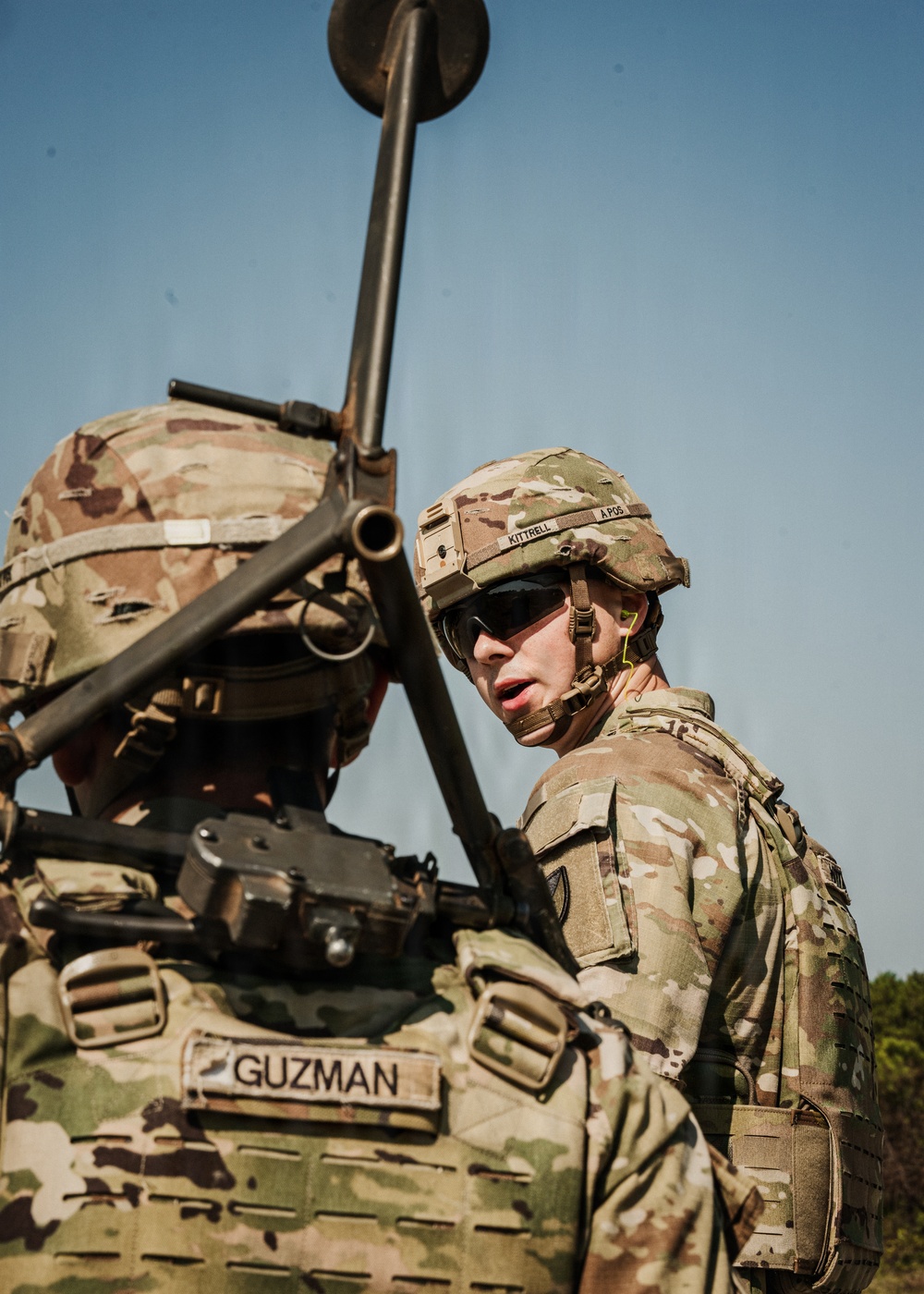 Two U.S. Army soldiers in full combat gear are engaged in conversation, likely during a field training exercise or operational briefing. The soldier facing the camera wears a helmet with name tape reading 'KITTRELL' and a blood type label 'A POS', while the soldier with their back to the camera has a name tape reading 'GUZMAN'. Both are equipped with OCP (Operational Camouflage Pattern) uniforms and tactical gear including body armor and communication equipment.