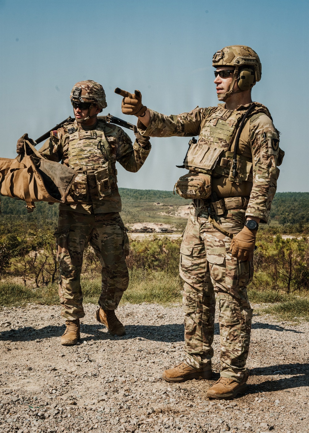 Two U.S. Army soldiers in full combat gear are standing on elevated gravel terrain, with one soldier pointing in a direction while the other carries equipment. Both are wearing Operational Camouflage Pattern (OCP) uniforms, plate carriers, and helmets. The background shows a wide open training range with vegetation and what appear to be military vehicles in the distance.